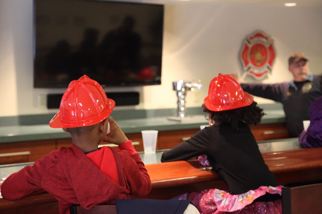A boy and a girl facing away from the screen sit at a Firehouse's bar drinking water while wearing plastic firefighter helmets.