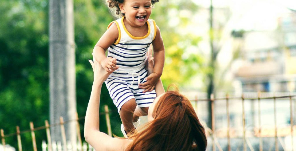 A woman, facing away from the screen, tosses a laughing child in the air. The child wears a white outfit with purple stripes and yellow shoulders.