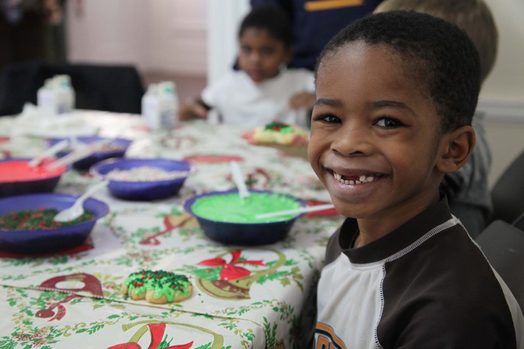 A boy missing his two front teeth smiles while sitting in front of a cookie decorating table