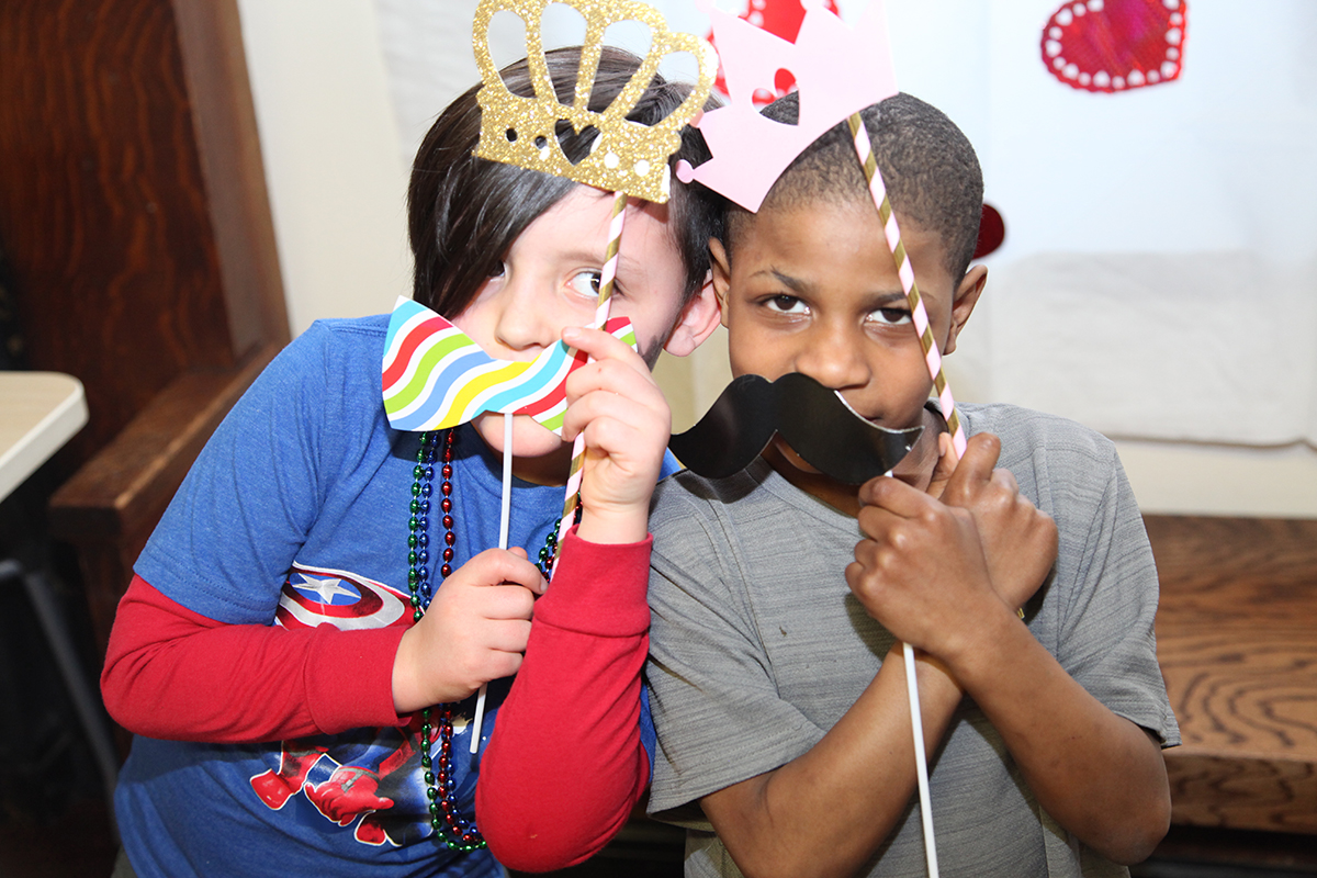 Two boys each hold photo booth props - a mustache and a crown - to their faces