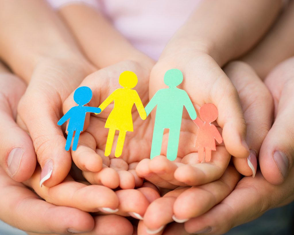 A man's hand holds a woman's hand, which holds a child's hand, which holds the cutout of a family of four, The family is blue, yellow, green, and red.