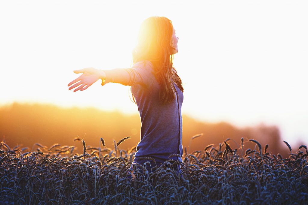 A woman stands in a field with her arms outstretched, facing away from the screen. The sunlight is bright.