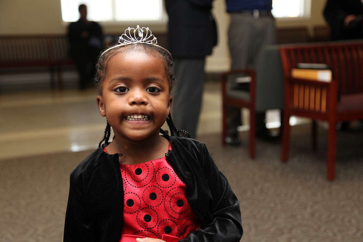 A young girl wearing a tiara, a red dress, and a black sweater smiles toward the camera.