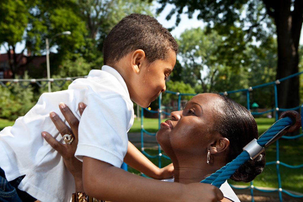 A woman holds up a young boy and looks at him with love. The boy is also holding on to a blue rope and has a few beads of sweat near his sideburn.