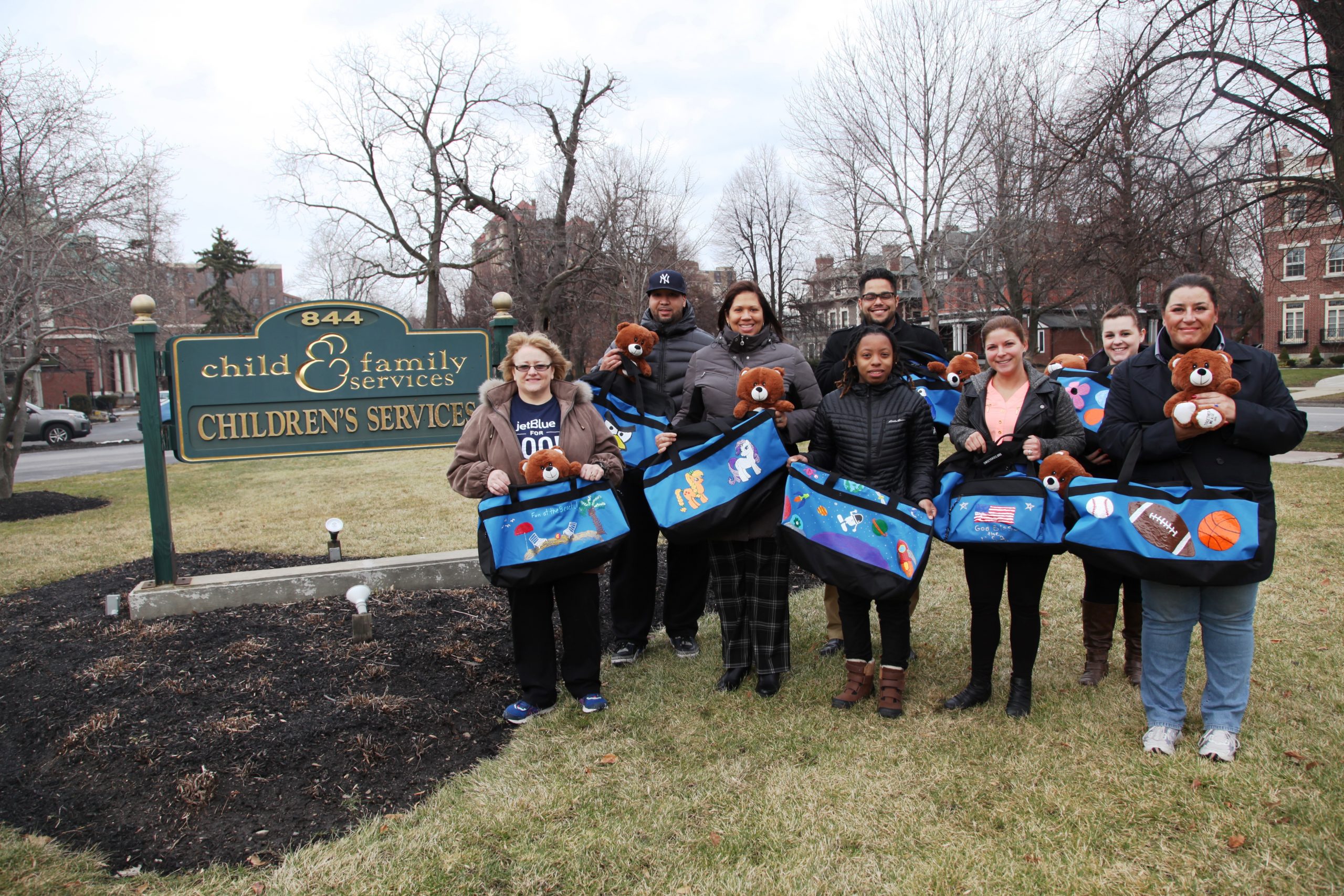 A group of eight hold blue duffel bags in front of the Child & Family Services lawn sign