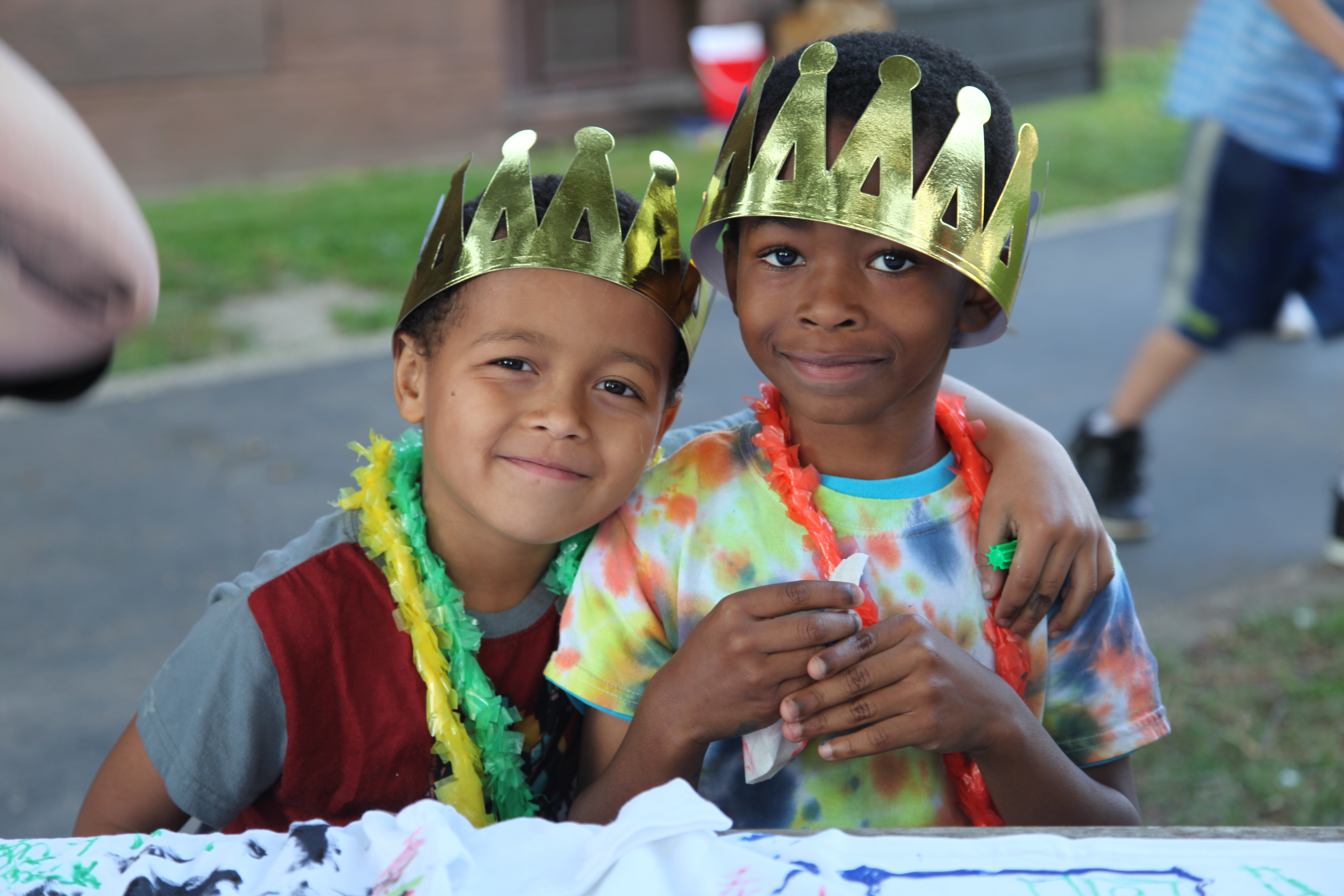 Two boys wearing crowns and leis. The one on the left has his arm around the one on the right.