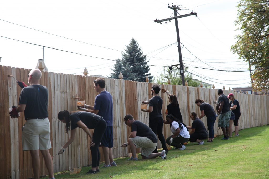 A group of twelve repaints a fence. Some are standing upright, some are crouched down.