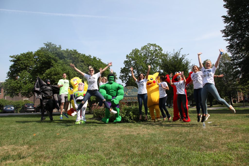 A group leaps for joy on a lawn. Some are dressed in costumes, including Batman, Big Bird, Buzz Lightyear, the Incredible Hulk, Pikachu, and Elmo.