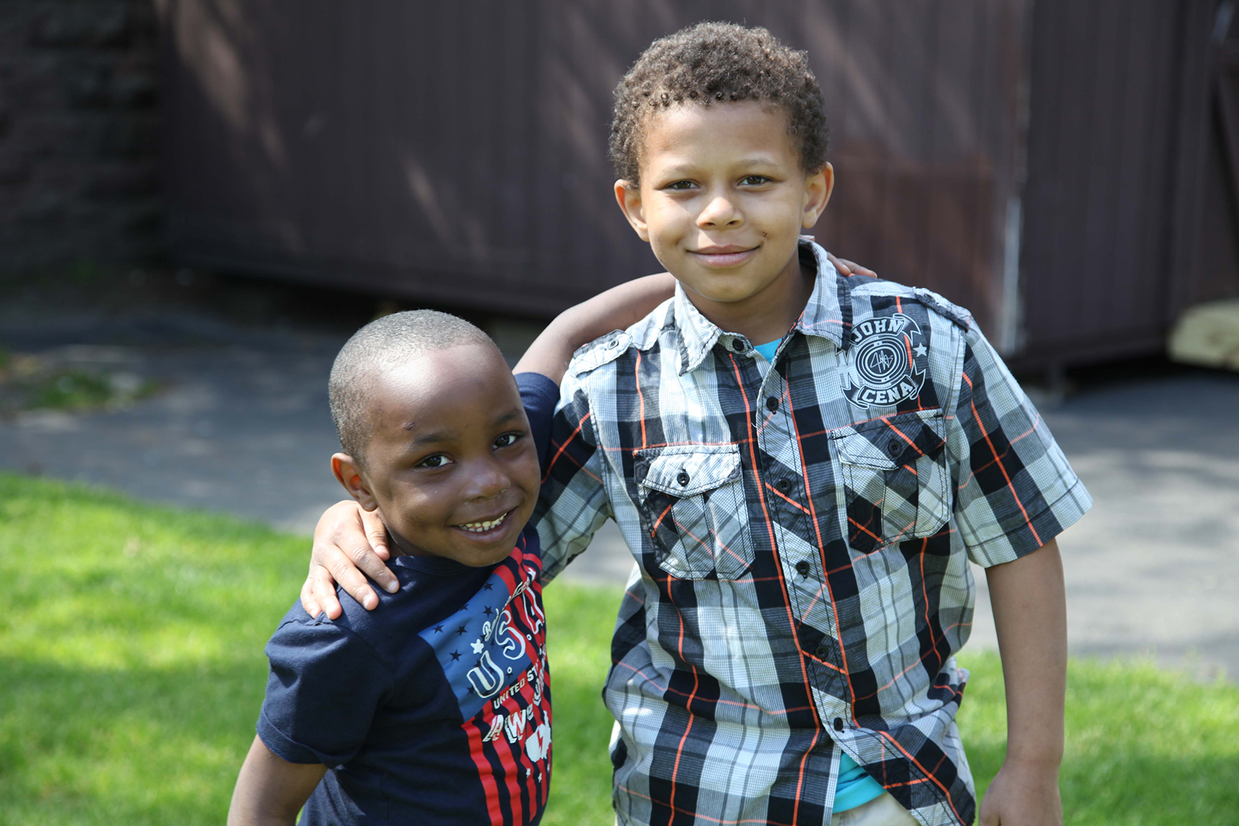 A small boy on the left and a taller boy on the right have their arms around each other's shoulder.