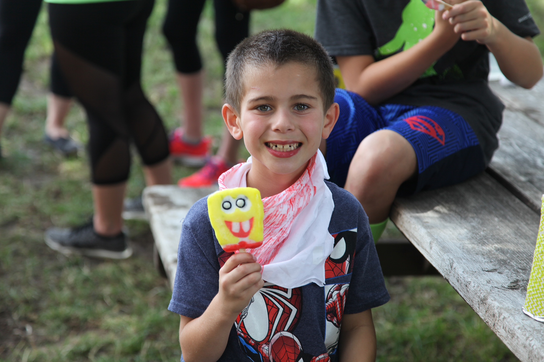 A boy wearing a Spider-Man shirt smiles toward the screen with a Spongebob popsicle in his right hand.