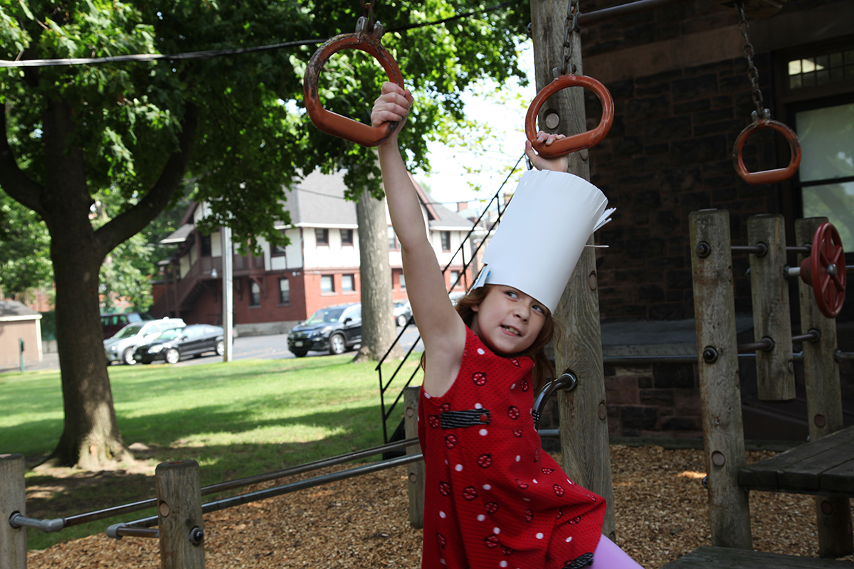 A young girl in a chef's hat and a red shirt climbs on a playground
