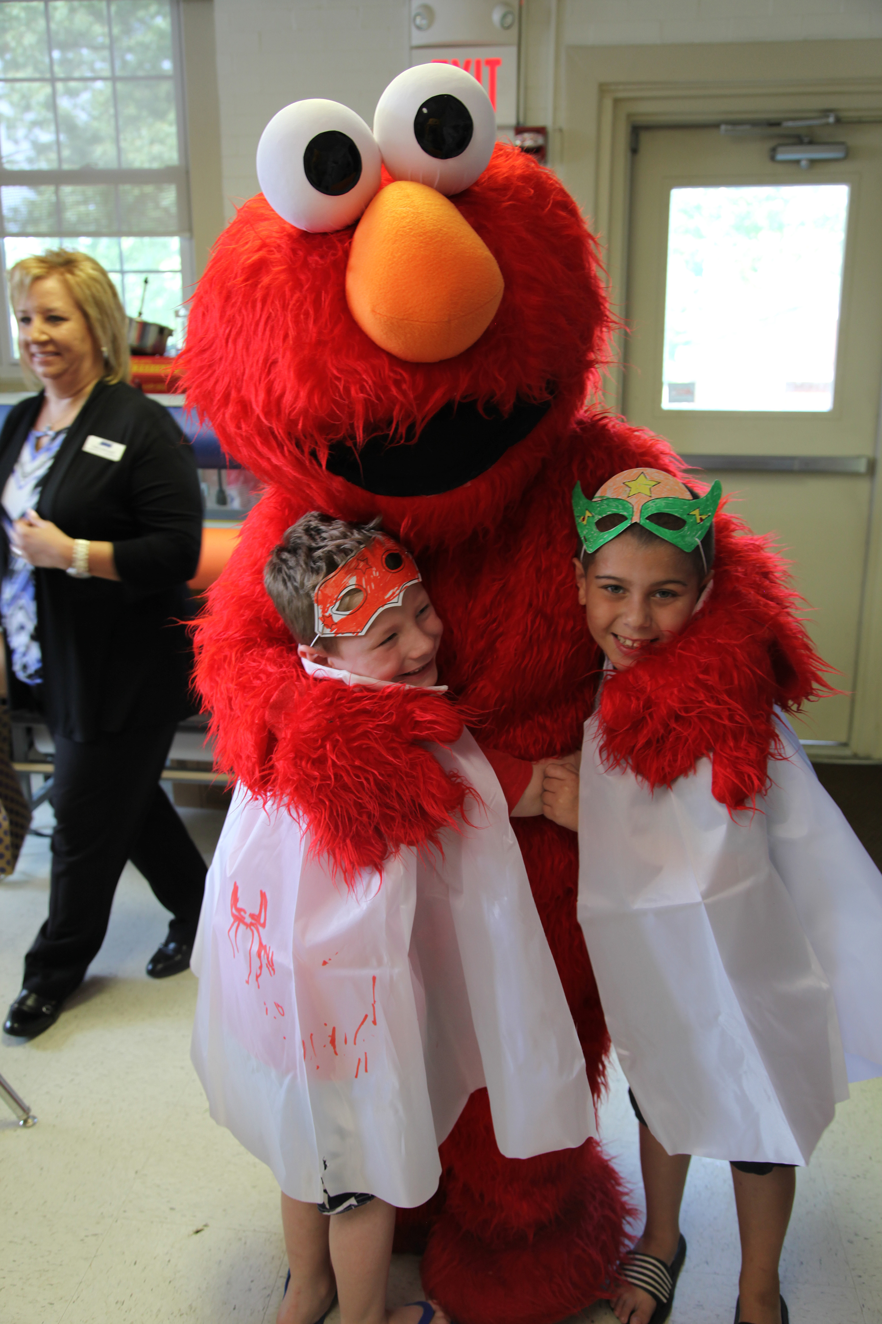 Two boys wearing protective cloths and masks are embraced by Elmo.