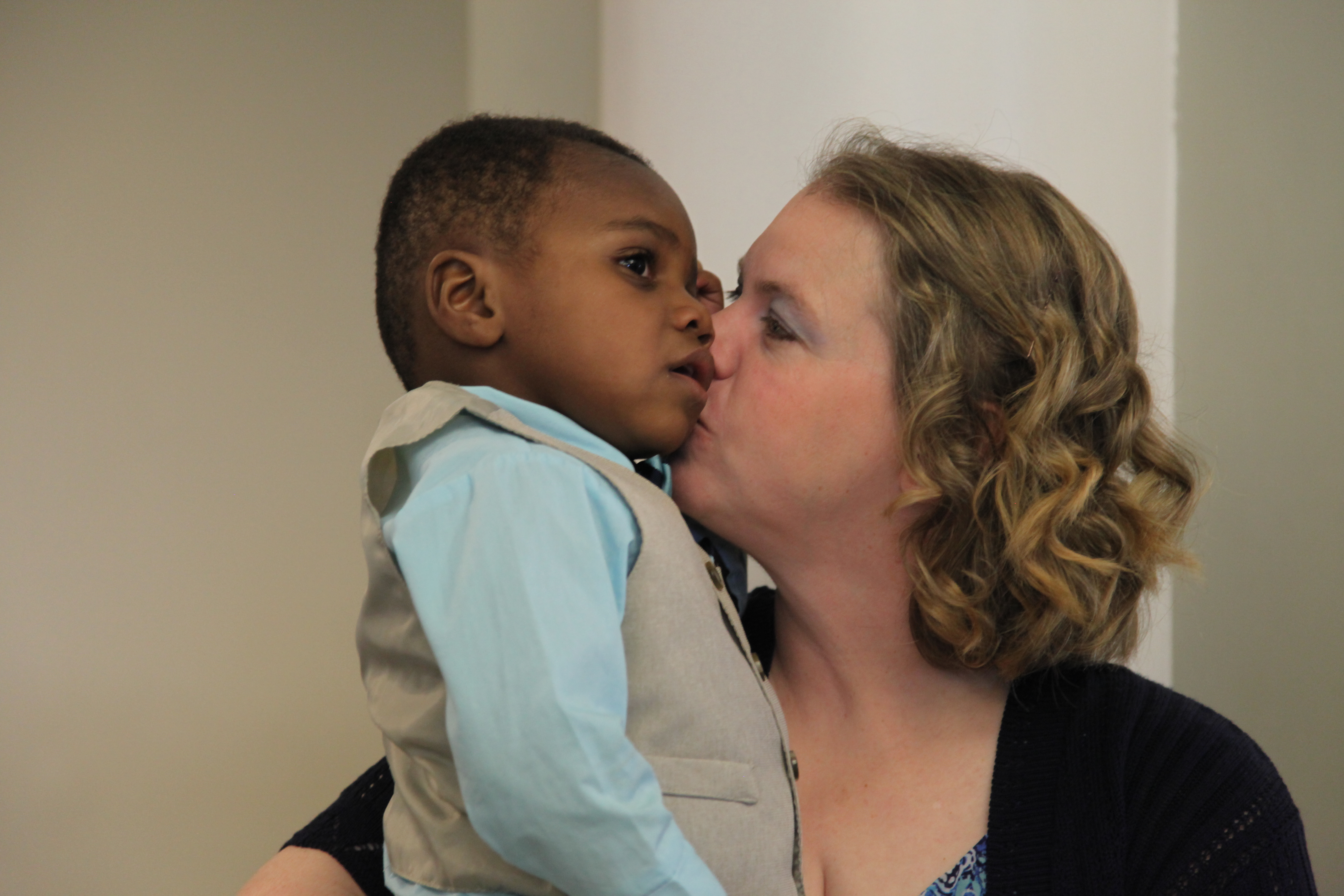 A white woman kisses the cheek of a young black boy she is holding. The boy is wearing a suit vest and a blue shirt.