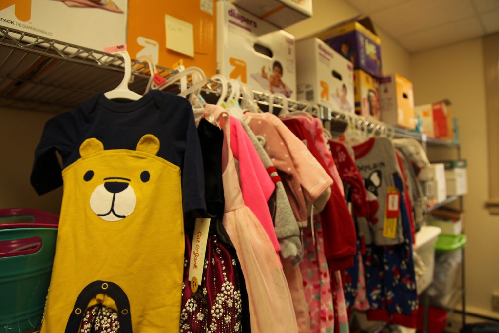 Onesies hang on the side of a rack that is holding diapers. The onesie in the front has a bear on it.