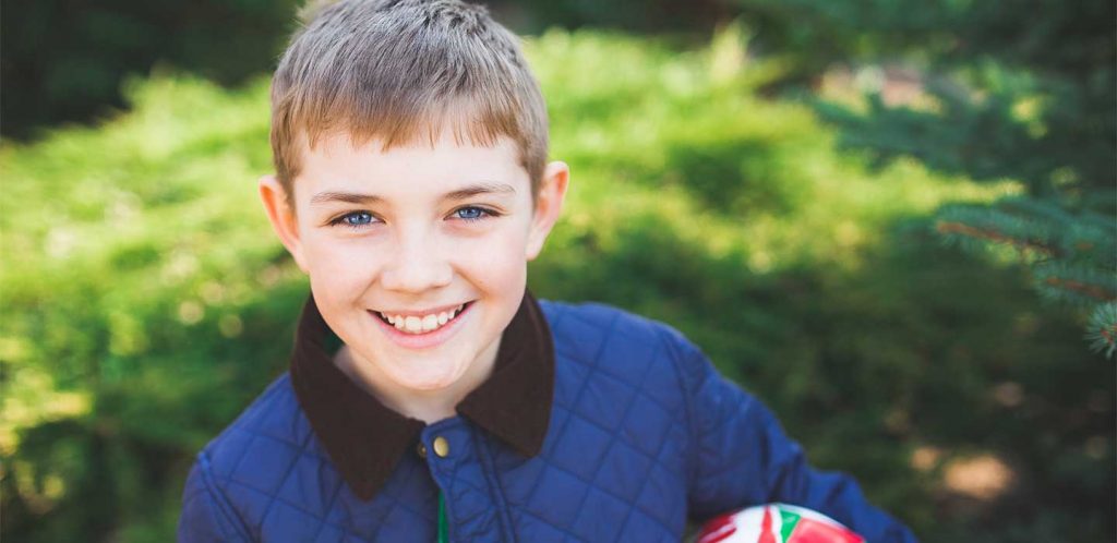A young boy with blue eyes and a blue jacket holds a soccer ball and smiles