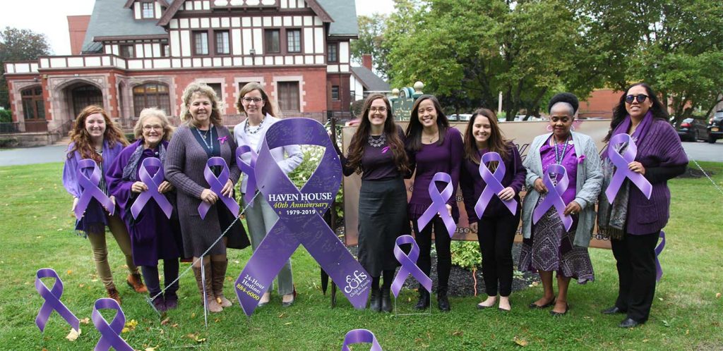 Nine women, all wearing purple, smile on a lawn while holding purple Domestic Violence Awareness banners. An oversized purple banner is in the middle of the group.