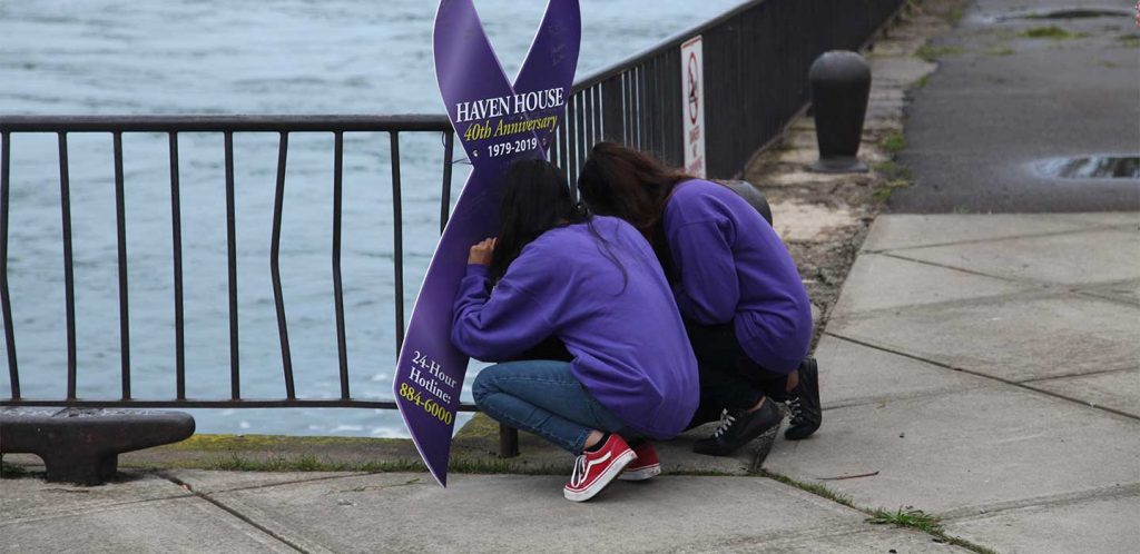 Two teenages, faces obstructed, wearing purple sign an oversized purple Domestic Violence Awareness Month banner cutout