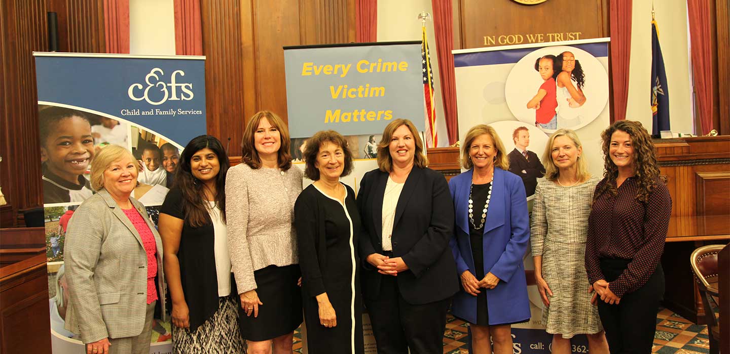Eight women smiling in front of a judge's bench and three vertical banners in a courtroom.
