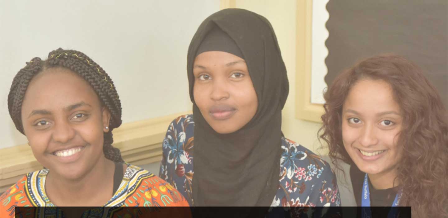 Three girls smile toward the camera. The girl in the middle is wearing a hijab.