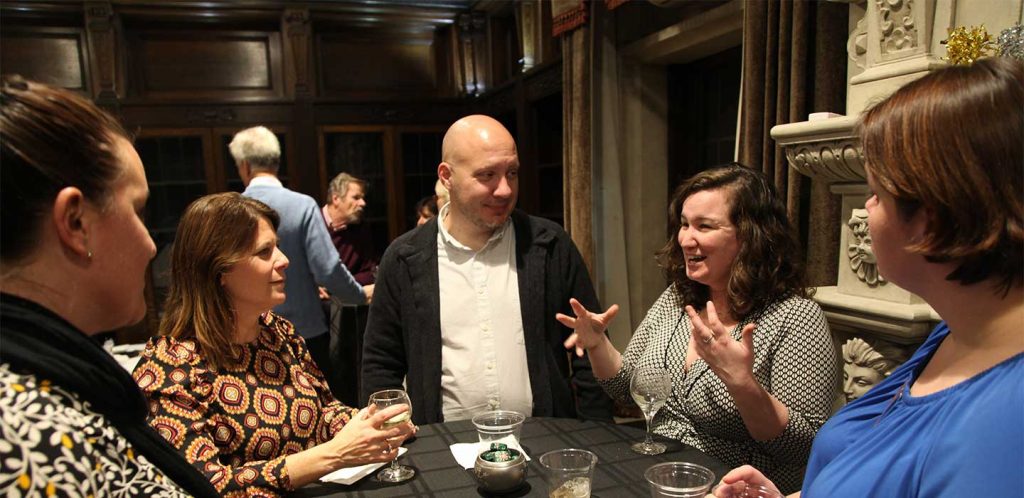 Three women and one man stand at a table with wine glasses listening to a fourth woman speak energetically, hands in the air