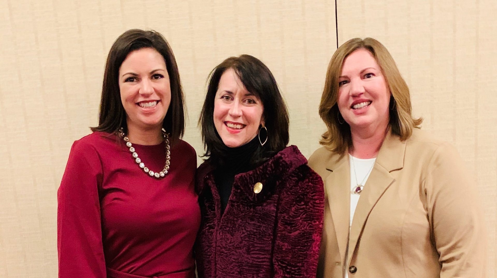 Three women stand in front of a blank wall smiling