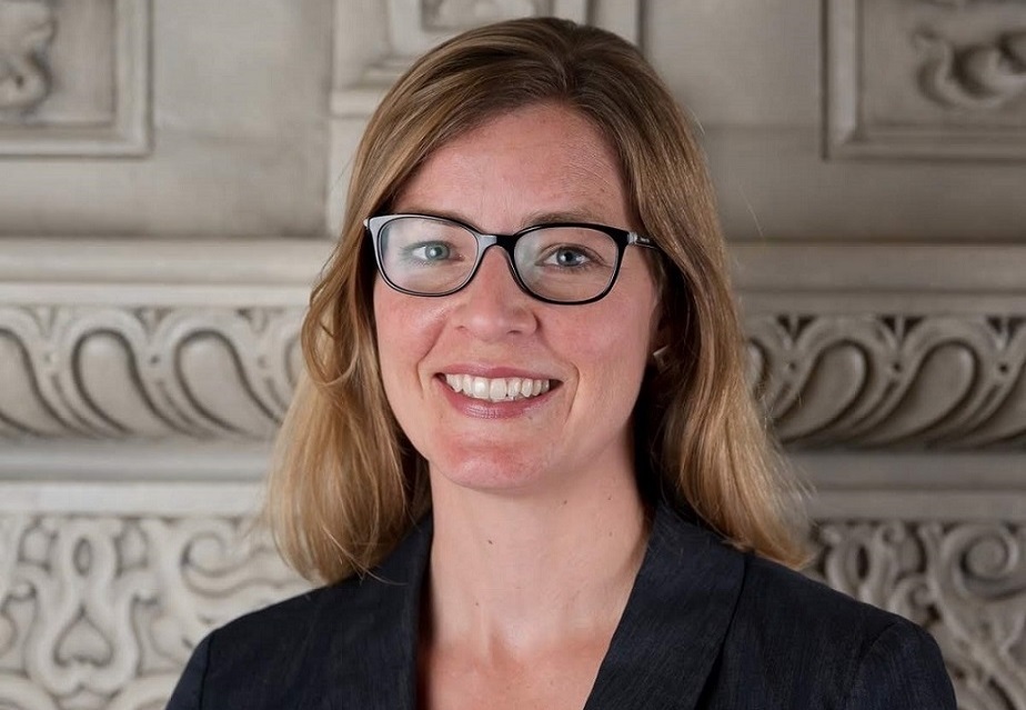 A professional woman wearing glasses and a suitcoat smiles to the camera