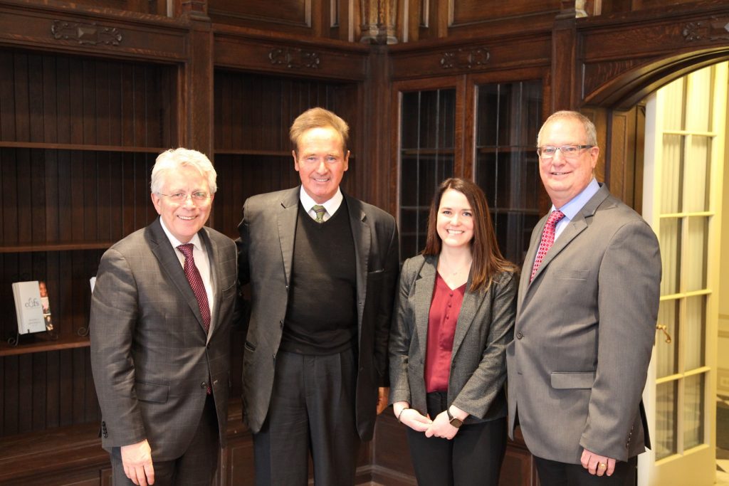 Three men and a woman dressed in business attire smile for a photo in front of wooden shelving