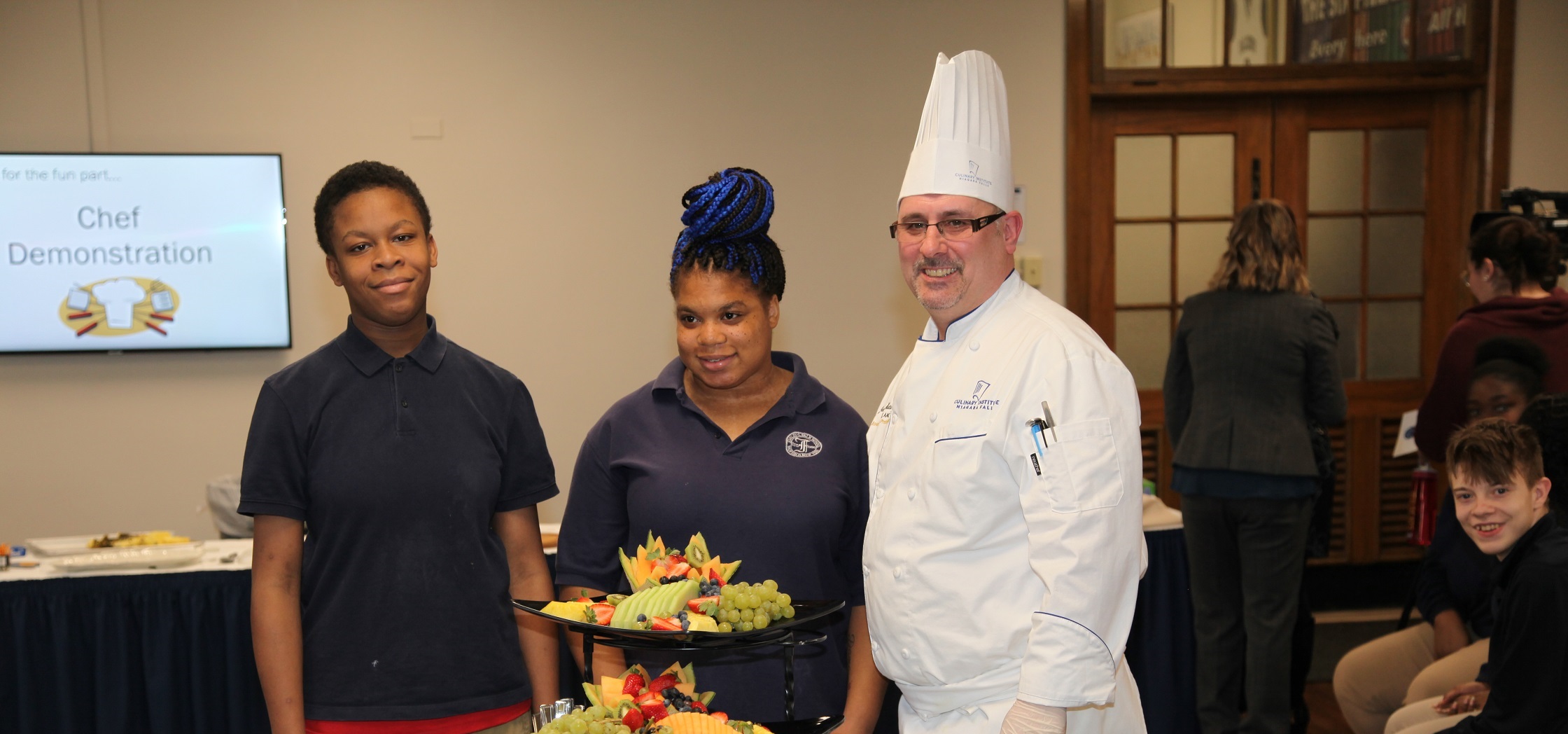 A male student, female student, and chef stand behind a display of fruit while smiling at the camera.