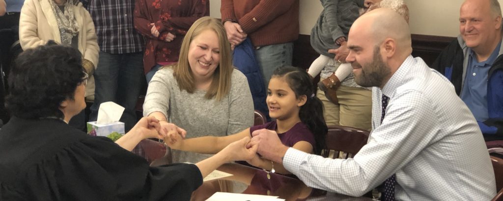 A woman, young girl, and man hold hands with a judge at the girl's adoption ceremony. All are smiling while seated at a wood table.