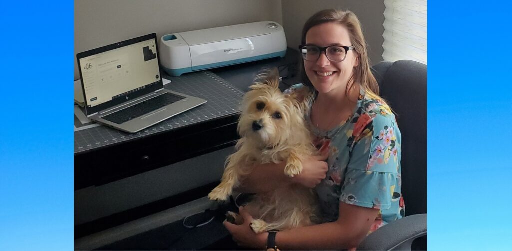 A woman wearing glasses holds her dog while sitting in front of her desk with a laptop