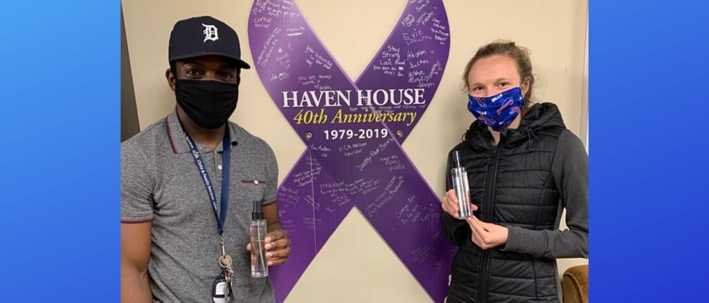 A man and woman wear masks and hold sanitizer while standing in front of a purple ribbon sign