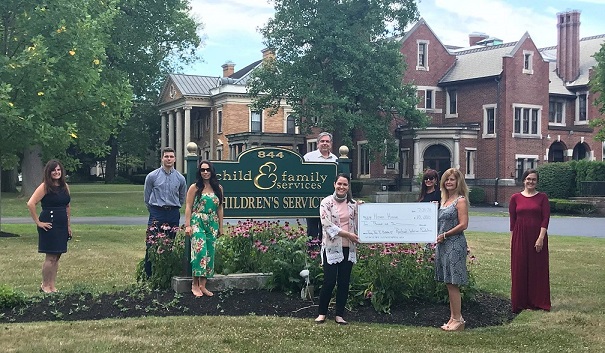 A group of people stand on a lawn. One woman hands a large check to another woman.