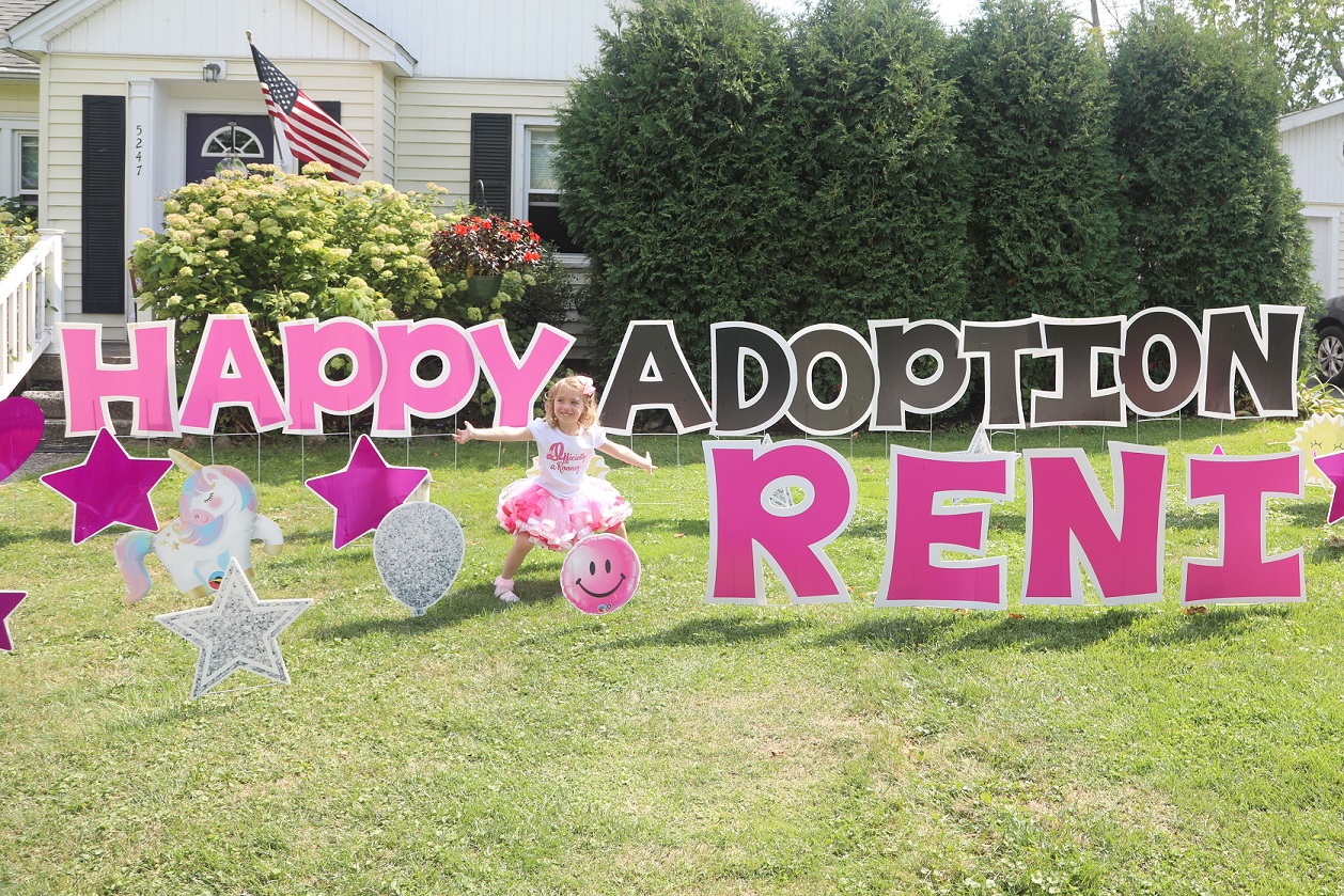 A young girl stands in front of a large lawn sign that reads "Happy Adoption Reni"