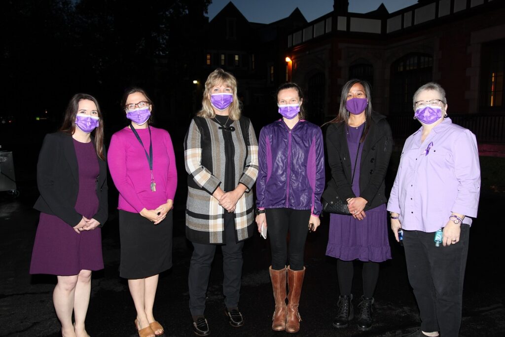 A group of six women wear purple masks outside at night
