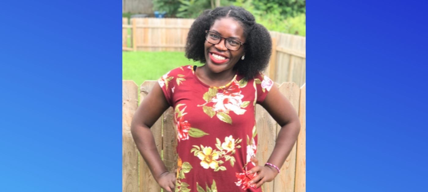 A young woman wearing a red, flowered dress stands outside with her hands on her hips and smiles at the camera