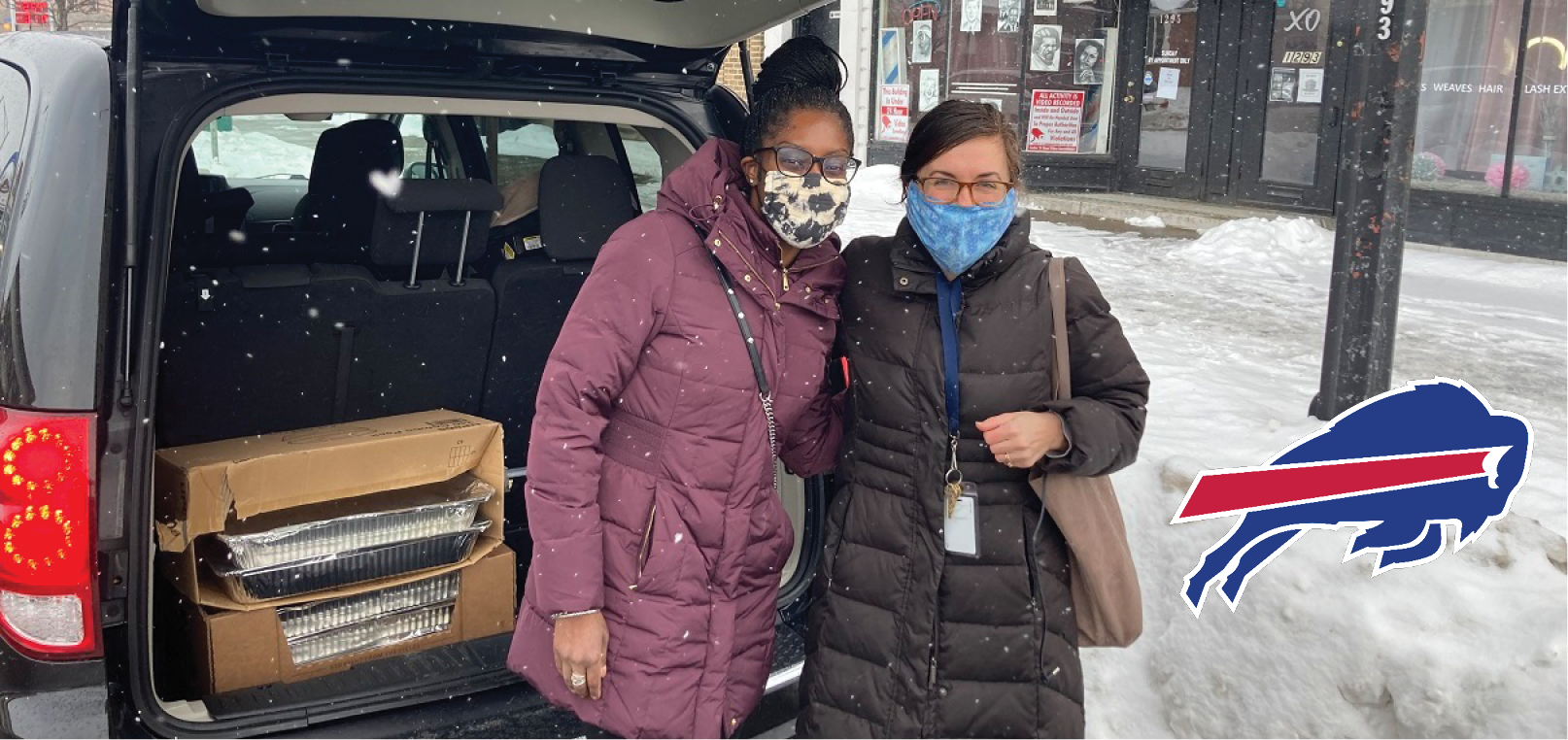 Two women wearing masks stand in the snow outside of a car's open trunk