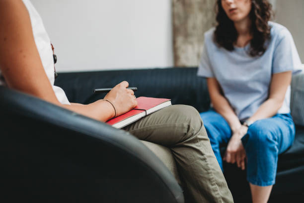 A teenage girl sits on a couch while a therapist sits across from her, listening. Both faces are out of frame.