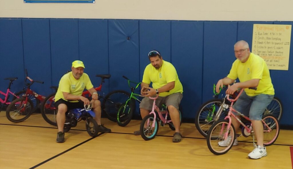 Three men in yellow shirts sit on bicycles inside a gymnasium