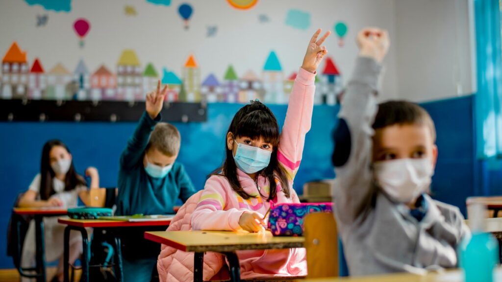 Students wearing masks at their school desks raise their hands