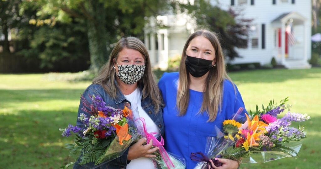 Two women wearing masks stand on a lawn outside while holding bouquets of flowers