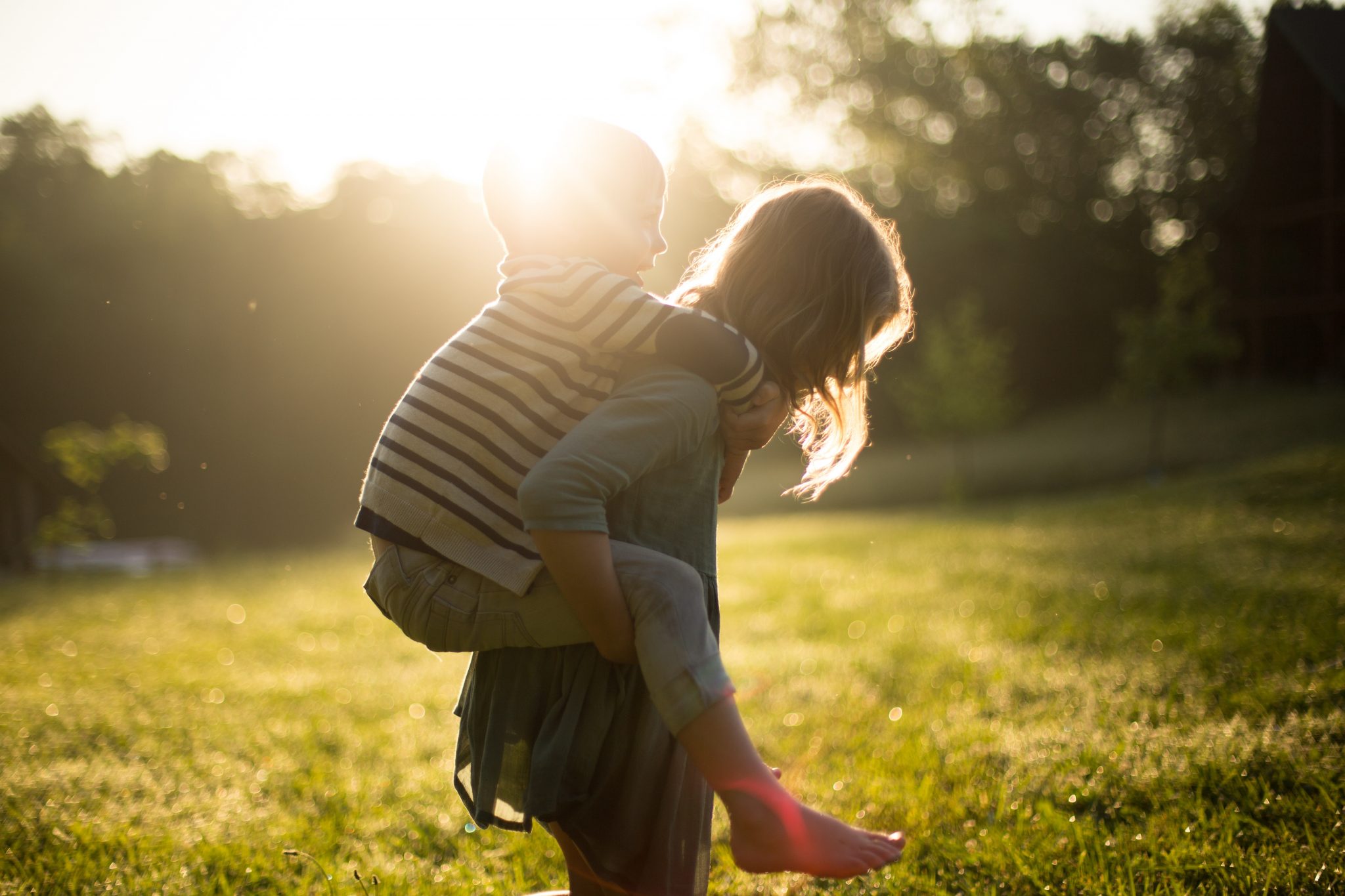 A girl holds her brother on her back in a field at sunset