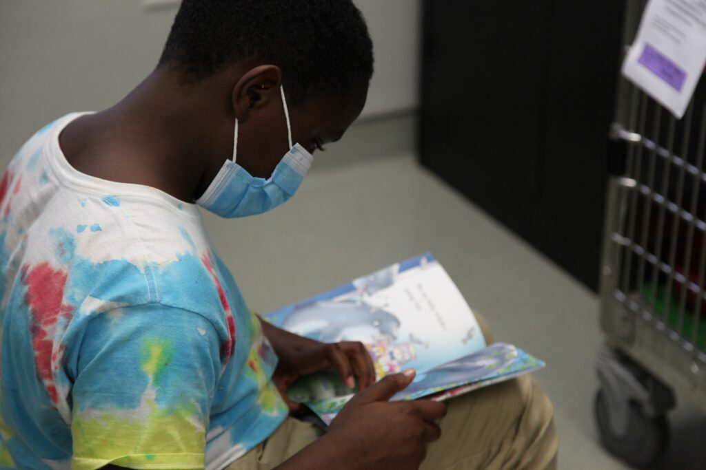 A boy in a multi-colored shirt and a mask reads a book in front of an animal cage