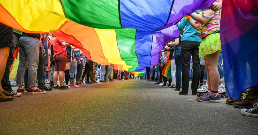 The underside of a rainbow flag being carried by people walking down a street.