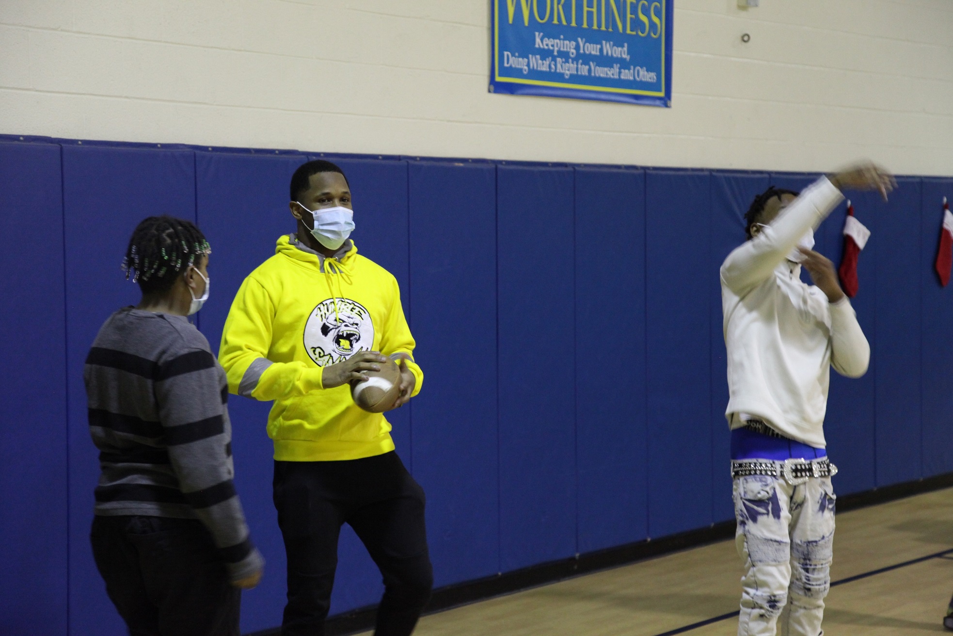 A man in a yellow hoodie speaks to a boy while another man throws a football behind them.