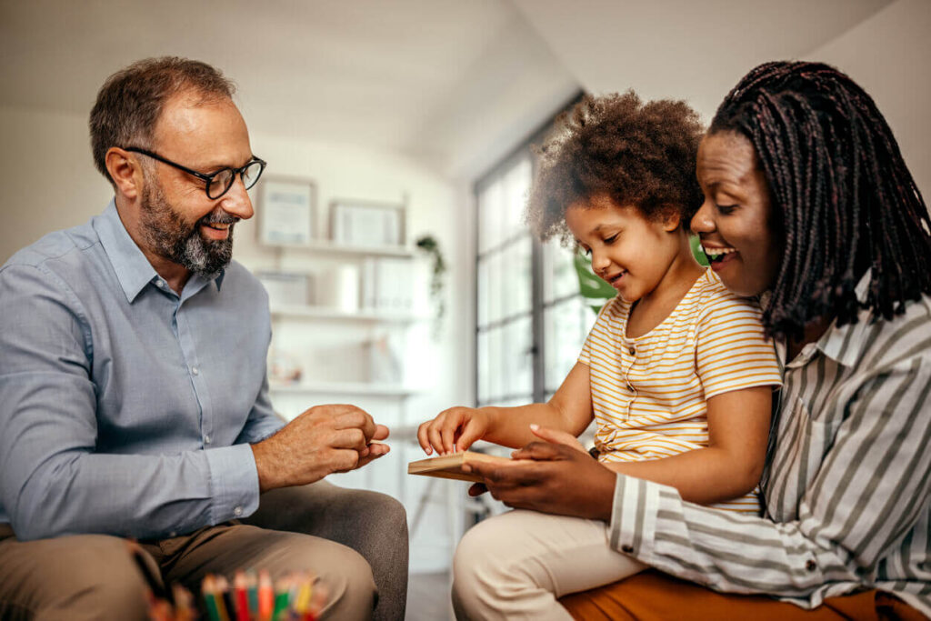 A child sits on their mother's lap. Both are smiling. The child is looking at a small activity board that the mother holds. A smiling counselor looks on.